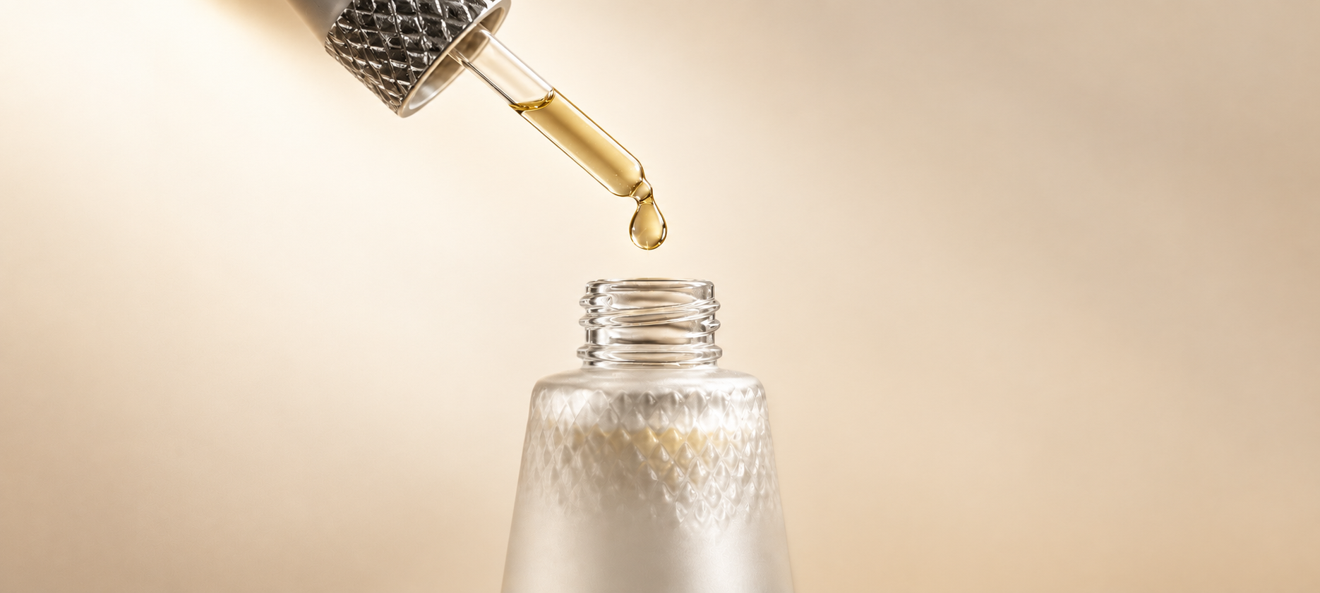 A golden drop of essential oil suspended above a frosted glass bottle against a soft white-to-beige background.